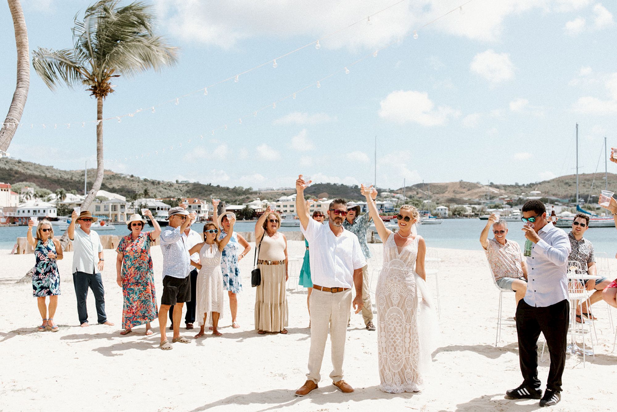 A group of people celebrates on a sunny beach with palm trees and scenic water views in the background.