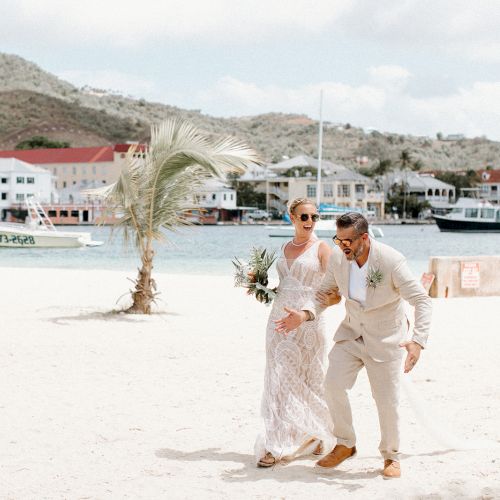 A couple in formal attire is playfully posing on a beach with boats and buildings in the background, under a partly cloudy sky.