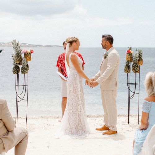 A couple is having a beach wedding ceremony, holding hands, with guests seated and pineapples as decor, set against an ocean backdrop.