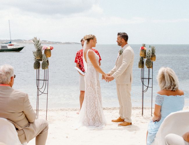 A couple is having a beach wedding ceremony, holding hands, with guests seated and pineapples as decor, set against an ocean backdrop.