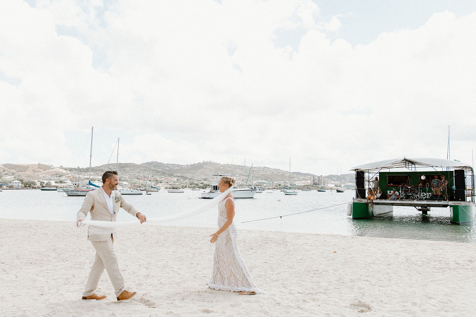 A couple in wedding attire walks on a beach near water, with a floating structure behind them.