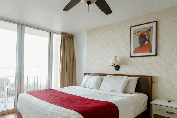 A neatly arranged hotel room with a large bed, red bedspread, side tables, and a ceiling fan, with a painting and window in the background.