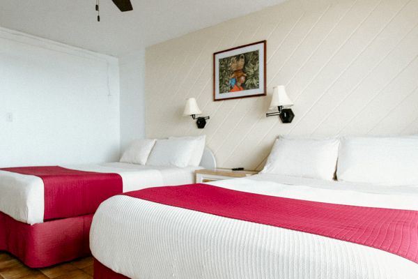 The image shows a hotel room with two beds, red bedspreads, a ceiling fan, and a framed artwork on the wall above the beds.