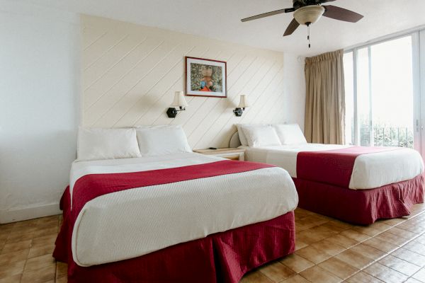 A hotel room with two beds, red bed skirts, wall lamps, a ceiling fan, and a window with curtains. A picture hangs above the beds.