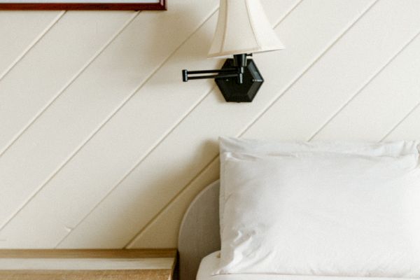 A cozy bedroom scene with a bed, a wooden nightstand, a wall-mounted lamp, and a framed artwork above the nightstand.