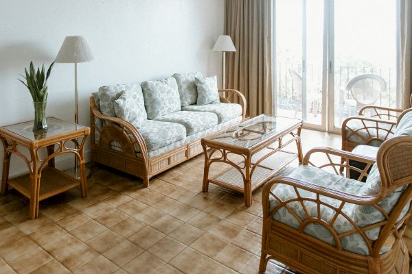 A living room with wicker furniture, cushioned seats, a glass-top table, lamps, and a plant, with tiled flooring and large windows.