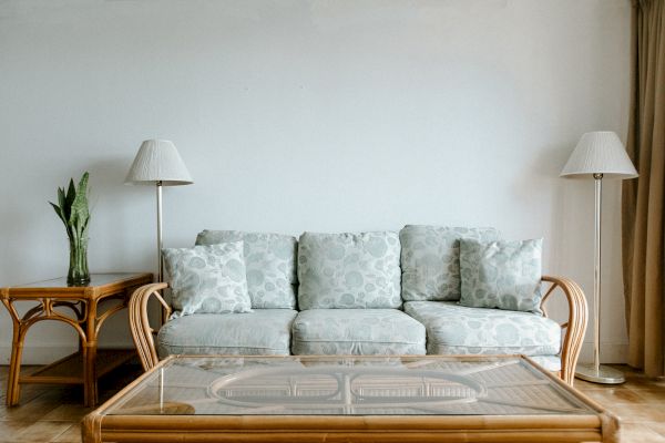 A living room with a sofa, glass coffee table, two lamps, and a plant on a side table against a plain wall.