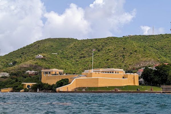 A coastal fort with yellow buildings sits by the sea against a green hillside under a blue sky with clouds.