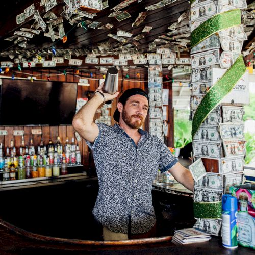 A bartender is shaking a mixer in a bar adorned with dollar bills pinned to the ceiling and walls.