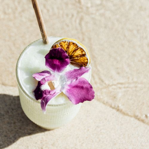 A refreshing cocktail with a purple flower garnish and a dried citrus slice, set on a sandy beach.