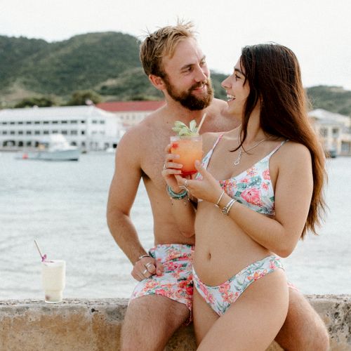 A couple in matching floral swimwear sits by the seaside, enjoying drinks and each other's company against a backdrop of mountains and water.