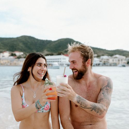 A woman and a man on a beach, smiling and toasting with drinks. The background shows water, boats, and hills.