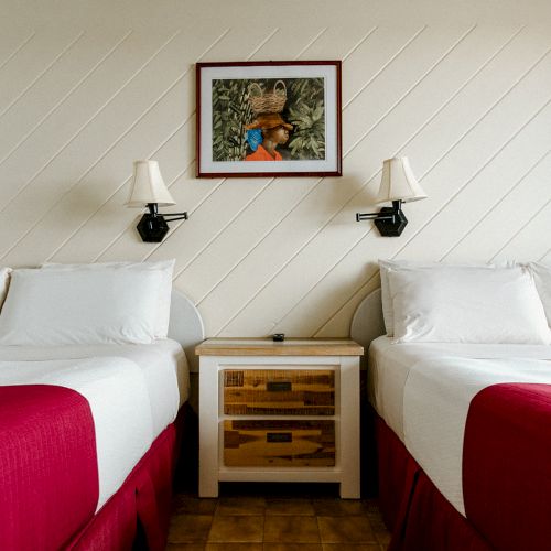 The image shows a hotel room with two double beds, white pillows, red bedspreads, a nightstand, wall lamps, and a framed artwork above.