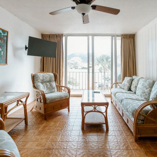 A cozy living room with wicker furniture, a ceiling fan, and a wall-mounted TV, overlooking a scenic view through large windows.