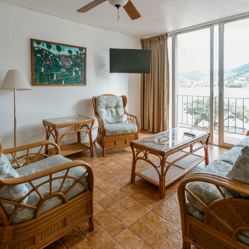 A cozy living room with wicker furniture, a table, a floor lamp, a painting, and a balcony view through large sliding doors.