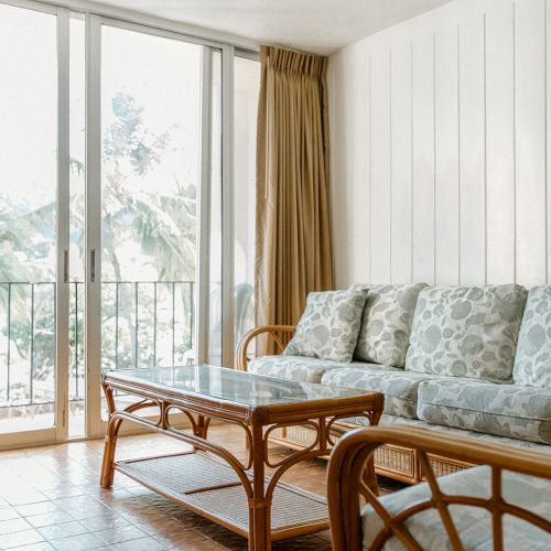 The image shows a bright living room with wicker furniture, a glass coffee table, sliding glass doors, and a view of greenery outside.