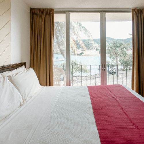 A hotel room with a neatly made bed, red runner, and a view of palm trees and water through glass doors with curtains.