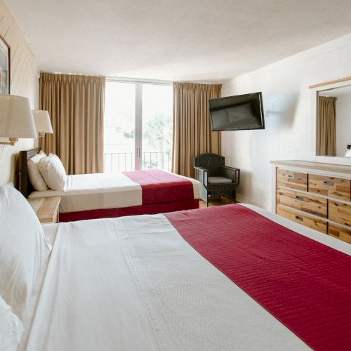 A hotel room with two beds, red accents, dresser, wall-mounted TV, chair, large mirror, and a window with curtains.