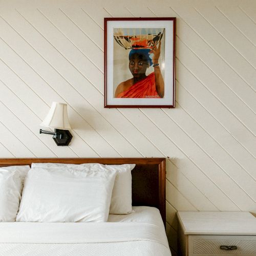 A neatly made bed with white pillows, a wall-mounted lamp, and a framed artwork on a diagonal wood-paneled wall.