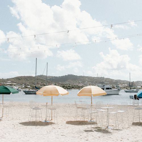 A sandy beach with colorful umbrellas, empty chairs, and a view of sailboats on the water under a blue sky with string lights above.