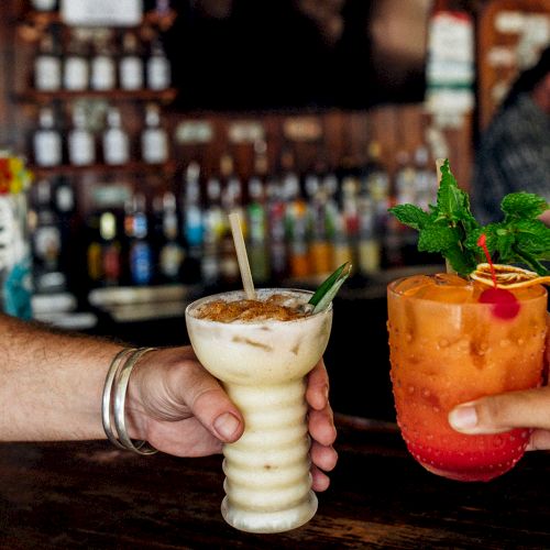 Two people holding colorful cocktails with garnishes in a bar setting, featuring bottles in the background and a person behind the counter.