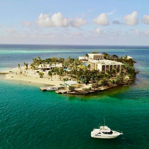 A small tropical island with sandy beaches, palm trees, a large building, and several boats in turquoise water under a partly cloudy sky.