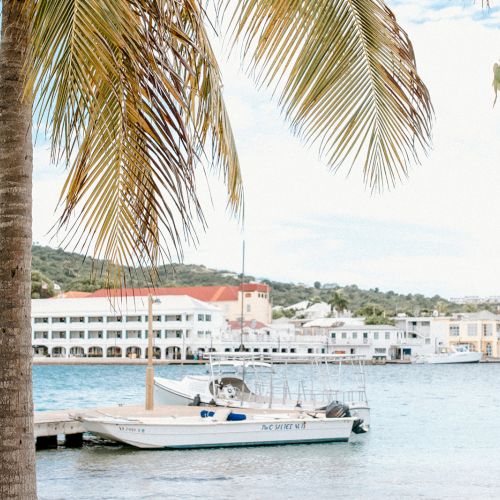 A calm harbor scene with a docked boat, palm tree, and buildings in the background under a blue sky ending the sentence.