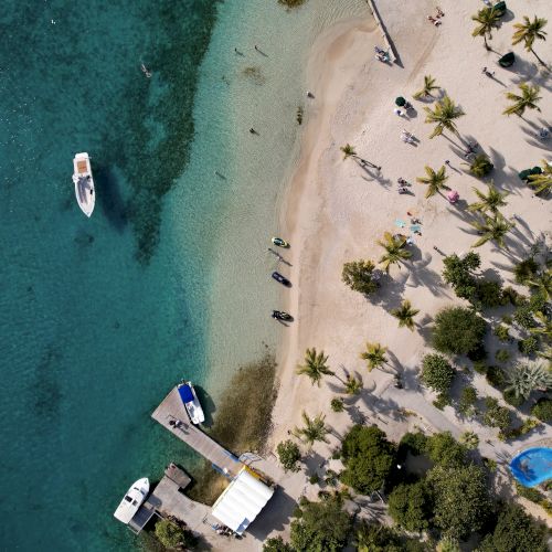 Aerial view of a tropical beach with palm trees, people, and boats in clear blue water. The scene captures a relaxing coastal vibe.