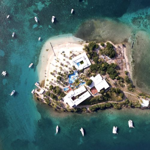 Aerial view of a small island with buildings, palm trees, and surrounding boats in turquoise water.